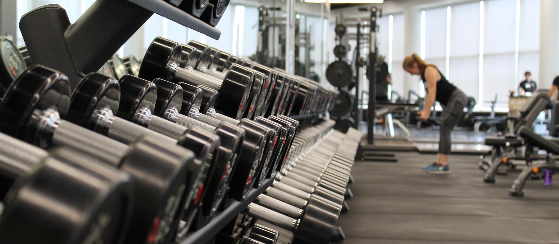 Rack med hantlar i gymmet och kvinna som tränar i bakgrunden.