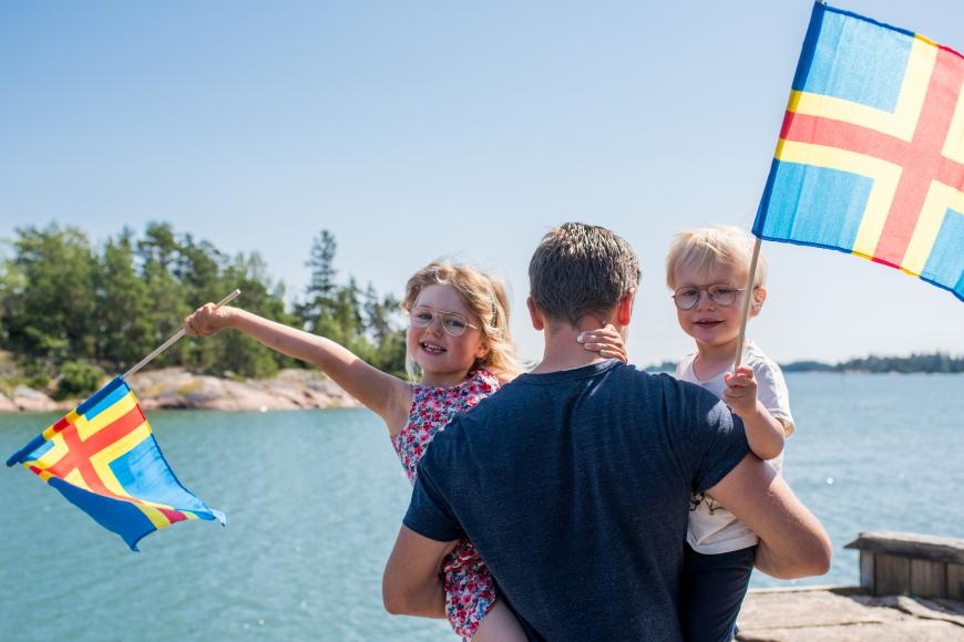 Två barn viftar med Ålandsflaggor i famnen på en vuxen.